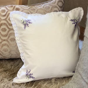 May include: A white square cushion with embroidered lavender sprigs and a delicate lace trim. The cushion has a soft, textured appearance and is likely for decorative use. The lavender embroidery adds a touch of floral elegance.
