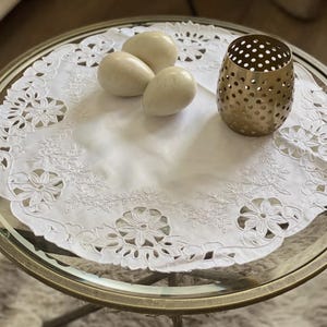 Peut inclure: Un napperon en crochet blanc avec un motif floral délicat est posé sur une table avec un plateau en verre et un piétement en métal doré. Trois œufs lisses et blancs et un bougeoir en métal doré sont placés sur le napperon.