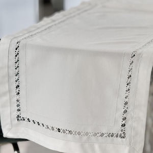 May include: A rectangular off-white table runner featuring a smooth central panel bordered by a wide hem. The border is decorated with an intricate, openwork cutwork embroidery pattern that creates a repeating geometric lattice design.