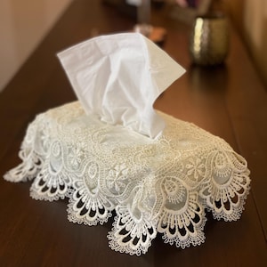 May include: White tissue box cover with delicate lace detailing. The cover is made of white lace and has a scalloped edge. The tissue box is not visible.