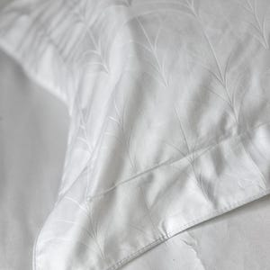 May include: A white pillowcase featuring a subtle, tonal jacquard pattern of repeating fan-shaped leaf motifs. The fabric has a smooth finish with a distinct border edge, showcasing a clean, monochromatic design across the entire surface.
