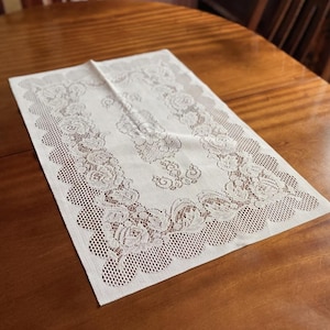 Könnte beinhalten: Cremefarbene rechteckige Spitzentischdecke mit Rosenmuster. Die Tischdecke liegt auf einem Holztisch. Die Ränder haben ein gewelltes Design.