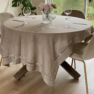Puede incluir: Un mantel redondo de lino beige con un borde decorativo, sobre una mesa de madera. La mesa está puesta con copas de vino, tenedores y cuchillos. Un jarrón de flores rosas está en el centro.