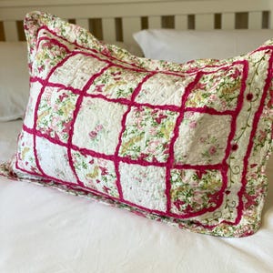 Op de afbeelding: Rechthoekig kussen met een bloemen patchwork ontwerp. Het kussen heeft een witte achtergrond met roze en groene bloempatronen, omlijst door een felroze rand. Het kussen ligt op een wit bed.