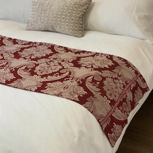May include: A bed runner featuring a deep red background with a repeating beige damask pattern. The runner is draped across a white bedspread, accompanied by a rectangular throw pillow with a beige and white wavy geometric pattern.