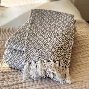 May include: A folded throw blanket featuring a repeating geometric diamond pattern in taupe and white. The fabric has a textured weave and is finished with long, cream-coloured fringe tassels along the bottom edge, creating a decorative border.