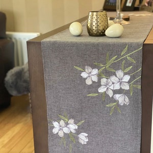 Könnte beinhalten: Ein grauer Tischläufer mit weißen gestickten Blumen und grünen Blättern. Der Läufer liegt auf einem Holztisch mit zwei weißen Deko-Eiern und einem goldenen Kerzenhalter.