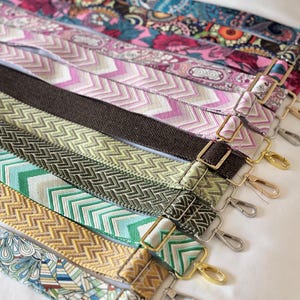 May include: A collection of adjustable bag straps featuring various patterns including colorful paisley, pink and white chevron, solid dark brown herringbone, olive green herringbone, teal and white chevron, and yellow chevron designs with metal hardware.