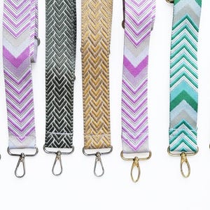 Pode incluir: Uma coleção de alças de bolsa coloridas e estampadas. As alças apresentam vários designs de chevron em tons de verde, roxo, dourado e cinza. Cada alça tem um fecho de metal para fácil fixação.