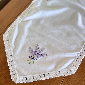 May include: A white rectangular table runner featuring embroidered purple lavender sprigs tied with a dark purple bow at each pointed end. The runner is finished with a delicate white lace trim along all edges, creating a decorative border.