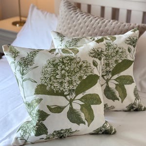 Puede incluir: Dos cojines cuadrados con un estampado botánico de flores de hortensia y hojas verdes sobre un fondo blanco roto. La tela muestra ilustraciones detalladas de racimos florales y follaje, creando un patrón consistente en ambos cojines.