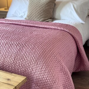 Può includere: Un copriletto trapuntato rosa antico con un motivo ovale ripetitivo e testurizzato drappeggiato su un letto. Il letto presenta due cuscini bianchi e un cuscino quadrato centrale beige con una trama a griglia, davanti a una testiera bianca.