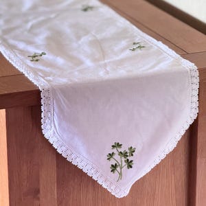 Op de afbeelding: Een witte rechthoekige tafelloper met geborduurde groene klavermotieven verspreid over de stof. De loper is afgewerkt met een delicate witte gehaakte kanten rand langs de zijkanten, wat zorgt voor een decoratieve afwerking van het textiel.