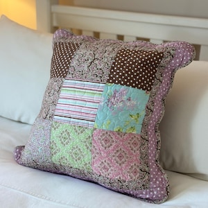 Op de afbeelding: Een patchwork kussen met diverse stoffen met patronen in bruin, roze, groen en blauw. Het kussen heeft een gerimpelde rand en ligt op een wit bed.