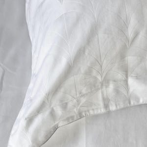 May include: A white pillowcase featuring a subtle, tonal jacquard pattern of repeating fan-shaped or leaf-like arches. The fabric has a smooth, soft texture with a slight sheen, and the design is woven directly into the white textile material.