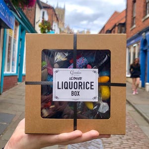 May include: A brown cardboard box filled with colourful sweets, visible through a clear window. The box is tied with black ribbon and has a label reading "Goodies Ultimate Liquorice Box". The box is held in front of a street scene.