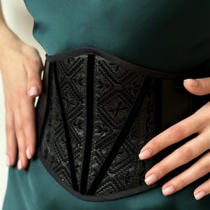 May include: A black corset belt featuring a central panel with an intricate embossed geometric pattern and star-like motifs. The belt is constructed with vertical velvet-textured piping accents and a smooth black fabric finish along the sides.