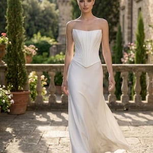 May include: A floor-length white gown featuring a structured, strapless corset bodice with vertical paneling and a smooth, flowing skirt. The garment is crafted from a solid white fabric, showcasing a clean, minimalist silhouette with a pointed hem.