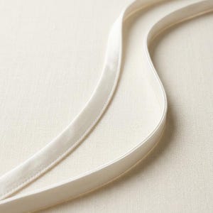 May include: Two long, smooth, off-white satin ribbons with finished stitched edges. The ribbons are arranged in gentle, flowing curves, showcasing a lustrous, reflective surface texture that catches the light across their length.