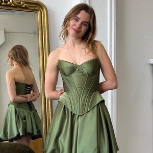 Peut inclure: Robe corset vert olive sans bretelles, avec un corsage structur&eacute; et une jupe &eacute;vas&eacute;e. La robe pr&eacute;sente une silhouette ajust&eacute;e et un aspect satin&eacute;. Le dos de la robe est visible dans un miroir, montrant un d&eacute;tail de la&ccedil;age. C'est une robe de soir&eacute;e.