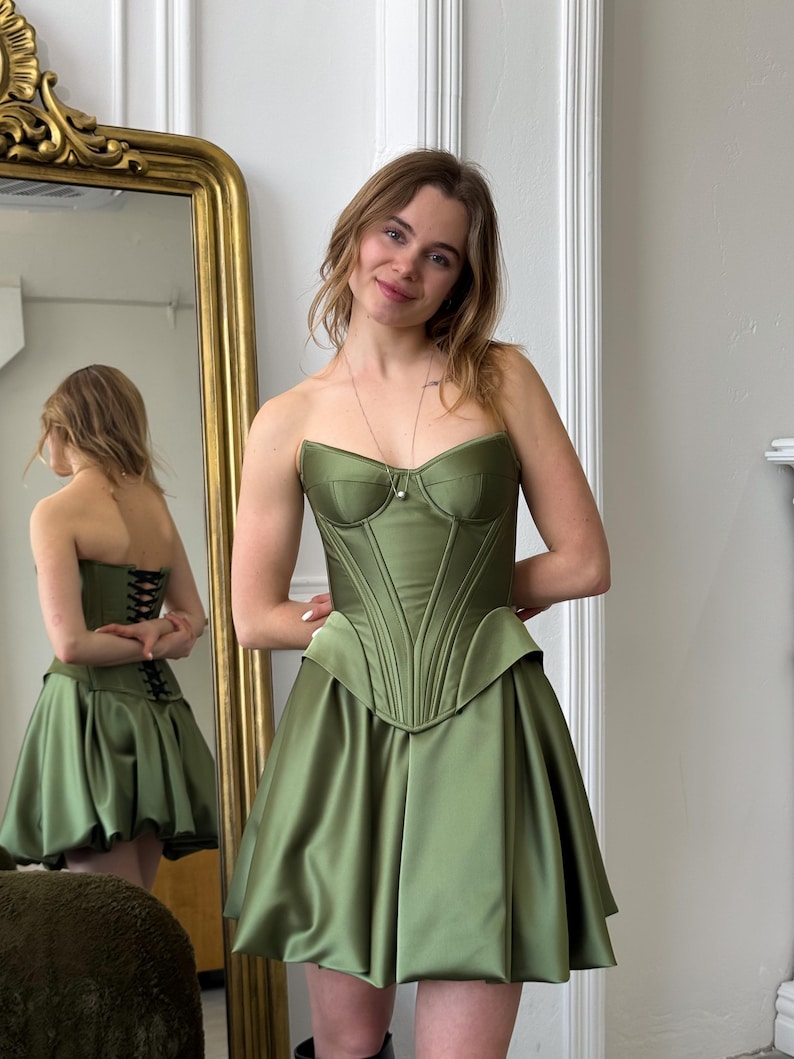 Peut inclure: Robe corset vert olive sans bretelles, avec un corsage structur&eacute; et une jupe &eacute;vas&eacute;e. La robe pr&eacute;sente une silhouette ajust&eacute;e et un aspect satin&eacute;. Le dos de la robe est visible dans un miroir, montrant un d&eacute;tail de la&ccedil;age. C'est une robe de soir&eacute;e.