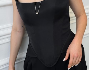 May include: Black strapless corset top with a structured design. The top has a curved neckline and a fitted silhouette. A silver necklace with a round pendant hangs from the neckline. The top is paired with black high-waisted pants.