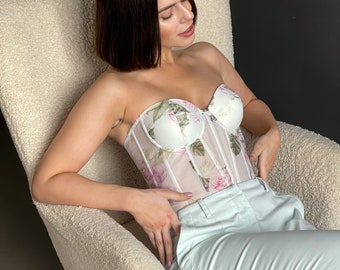 May include: A woman wearing a white floral corset with a sheer overlay and light blue pants, sitting in a white fluffy chair.