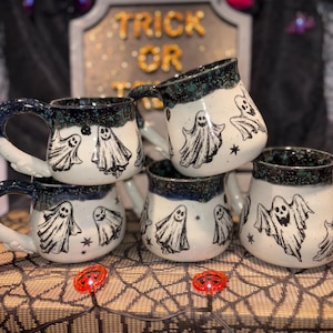 May include: A stack of white ceramic mugs with black ghost illustrations and star accents. The mugs have dark blue and green speckled rims and handles. A Halloween-themed display with a "Trick or Treat" sign is in the background.