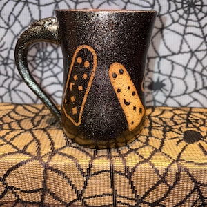 May include: A black ceramic mug with a textured, glittery finish. The mug features two orange, ghost-shaped designs with dot details. The handle has a metallic, iridescent finish. The mug is likely for Halloween or spooky season.