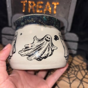 May include: A white ceramic Halloween mug with black ghost illustrations. The mug has a dark blue and gold speckled rim. A black skull and star are also featured on the mug. The mug is held in a hand.