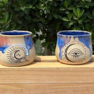 May include: Two ceramic mugs with celestial designs. One mug features a sun, moon, and stars, while the other displays a crescent moon with an eye. Both have blue and red glaze drips over a tan base.