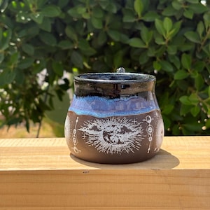 May include: A handcrafted ceramic jar with a celestial design. The jar features a dark brown base with a white illustration of the sun, moon, and stars. The rim is black, transitioning to a blue glaze. The jar is sitting on a wooden surface.