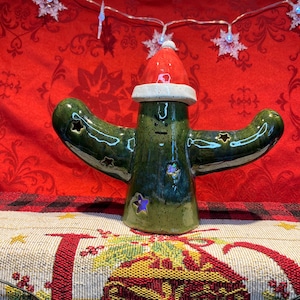 May include: A green ceramic cactus wearing a red Santa hat with white trim. The cactus has a face and is decorated with green and blue glaze. The cactus is sitting on a white and red patterned surface.