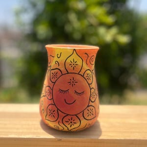 May include: Hand-painted ceramic vase with a smiling sun design. The vase features a gradient of yellow and orange hues, with black outlines and details. The sun's face has closed eyes and a gentle smile, adding a whimsical touch to the decorative piece.