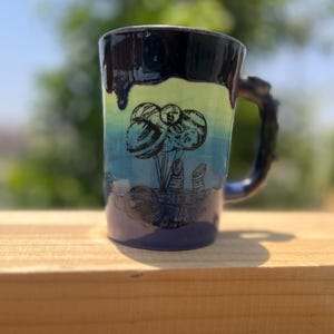 May include: A handmade ceramic mug with a unique handle and a colorful design. The mug features a dark blue rim that drips down the sides, transitioning to a light green and blue gradient. The mug has a black illustration of planets.