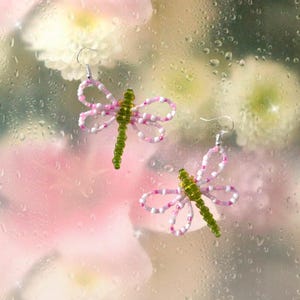 May include: A pair of dragonfly earrings. The earrings have green beaded bodies and wings made of pink and white beads. They hang from silver-coloured hooks, set against a soft, blurred background of pink and white.