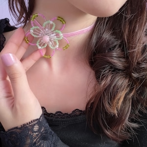 May include: A pink beaded flower choker necklace with a pink velvet ribbon. The flower has a central pink bead, surrounded by white beads, pink and green beaded petals. The necklace is worn around the neck.