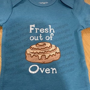 Könnte beinhalten: Ein türkisfarbener Baby-Body mit dem weißen Schriftzug "Fresh out of Oven" und einer Cartoon-Zimtschnecke. Der Body besteht aus weichem Stoff und hat kurze Ärmel.