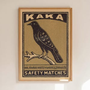 May include: A framed rectangular print featuring a black bird perched on a branch against a muted olive background. The text KAKA appears at the top, with KALISWARI MATCHWORKS SIVAKASI and SAFETY MATCHES printed in black at the bottom of the design.