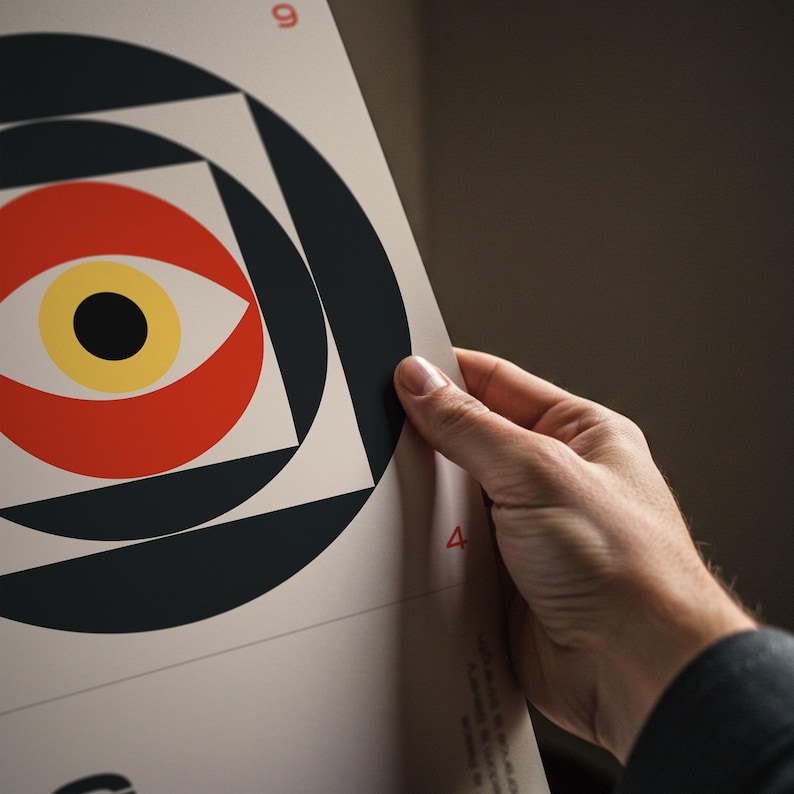 May include: Geometric art print featuring a stylized eye design. The artwork uses a palette of red, yellow, black, and white, arranged in circular and square shapes. The print is held by a person.