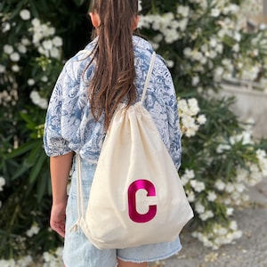 Può includere: Uno zaino a sacca in tela color crema con una grande lettera C di colore rosa acceso metallizzato stampata sul davanti. La borsa è dotata di spessi cordini bianchi come spallacci ed è realizzata in un tessuto semplice e resistente.