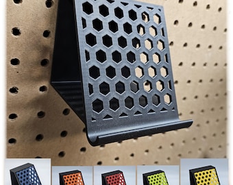 Pode incluir: Um suporte de telefone preto com um padrão de favo de mel. O suporte é feito de plástico e tem uma pequena borda na frente para segurar um telefone no lugar.