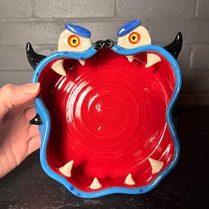 May include: A ceramic monster bowl with a vibrant red interior and a blue rim. The bowl features white teeth, black horns, and cartoonish eyes with yellow irises and blue caps. A unique and playful decorative piece.
