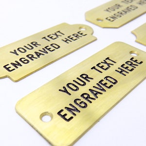 May include: Four gold-colored metal tags with rounded corners and holes. Each tag has the text "YOUR TEXT ENGRAVED HERE" engraved on the surface.