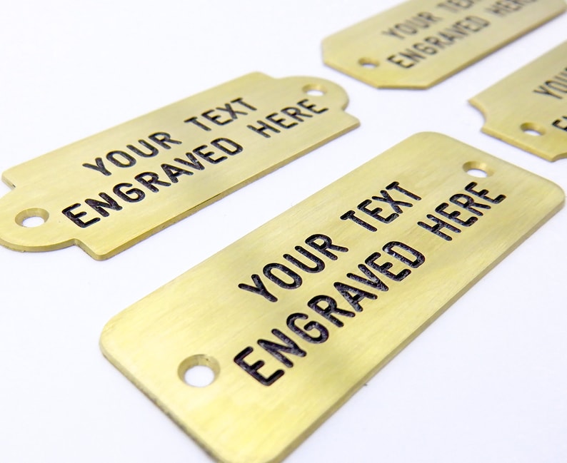 May include: Four gold-colored metal tags with rounded corners and holes. Each tag has the text "YOUR TEXT ENGRAVED HERE" engraved on the surface.