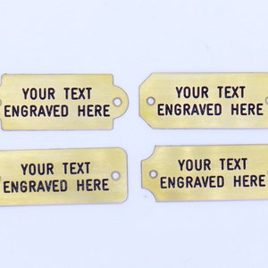 May include: Four gold-colored metal tags with rounded corners and holes. Each tag has the text "YOUR TEXT ENGRAVED HERE" in black.