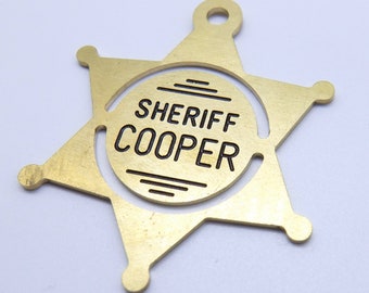 May include: A gold-colored sheriff's badge in the shape of a six-pointed star. The badge has the words "SHERIFF COOPER" in black lettering within a circular center.