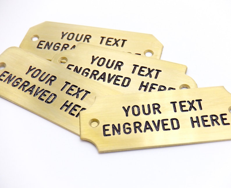 May include: Four brass tags with rounded corners and two holes. The tags are stacked on top of each other. The text "YOUR TEXT ENGRAVED HERE" is engraved on each tag.