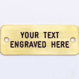 May include: A gold-colored rectangular metal tag with two holes. The tag has the text "YOUR TEXT ENGRAVED HERE" engraved on it.