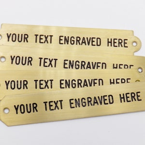 Può includere: Quattro etichette in ottone con angoli arrotondati e fori per l'appeso. Ogni etichetta ha il testo "YOUR TEXT ENGRAVED HERE" inciso in nero.